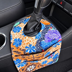 May include: A fabric car gear shift cover featuring a vibrant floral pattern in shades of blue, orange, and black. The cover includes a circular cutout for the gear shifter and a small integrated pocket on the front surface for storage.