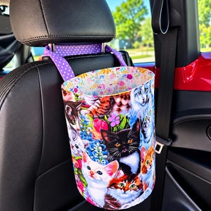 May include: A cylindrical fabric car trash bag featuring a colorful print of various kittens and flowers. The bag is suspended from a car headrest by two purple straps with white polka dots. The interior lining has a light floral pattern.