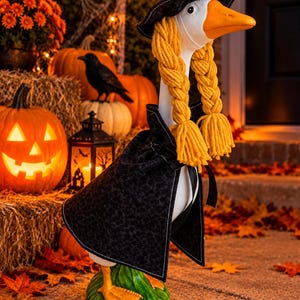 May include: A white goose figurine dressed as a witch, wearing a black patterned hat and cape. The goose has long, braided yellow yarn hair hanging down both sides of its head. It stands on a green base with orange webbed feet.