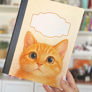 Puede incluir: Un cuaderno con una cubierta beige claro que presenta una ilustración detallada de la cara de un gato atigrado naranja. El cuaderno tiene un lomo negro y un área de etiqueta en blanco. El gato tiene ojos grandes y expresivos.