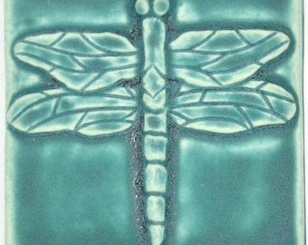 May include: A square ceramic tile in a teal glaze featuring a raised, embossed dragonfly design. The dragonfly has detailed wings with visible veins and a segmented body, with the relief areas highlighted in a lighter shade of teal.