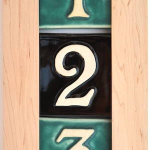 May include: A rectangular house number plaque with the numbers 1, 2, and 3. The numbers are cream-colored and set against a teal and black background. The plaque is framed in light-colored wood.