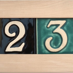 May include: A rectangular house number sign featuring the numbers 1, 2, 3, and 4. The numbers are in a cream color and are set against teal and navy blue tiles. The tiles are framed by a light-colored wooden border.
