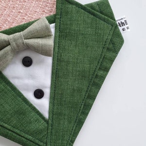 Może przedstawiać: Zielony materiałowy śliniak z białym przodem w stylu smokingu, muszką i dwoma czarnymi guzikami. Śliniak ma etykietę z napisem "Hand made in the"