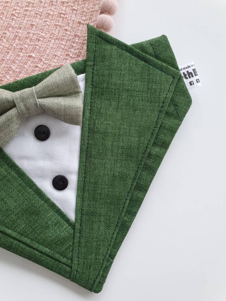 Może przedstawiać: Zielony materiałowy śliniak z białym przodem w stylu smokingu, muszką i dwoma czarnymi guzikami. Śliniak ma etykietę z napisem "Hand made in the"