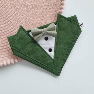 Może przedstawiać: Zielony kołnierz dla psa z tkaniny z białą muszką w stylu smokingu i dwoma czarnymi guzikami. Kołnierz ma napis "the handmade co." z tyłu.