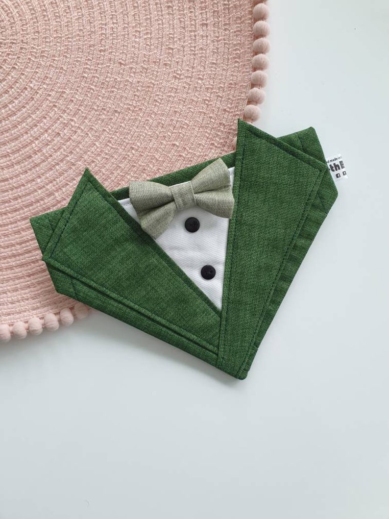 Może przedstawiać: Zielony kołnierz dla psa z tkaniny z białą muszką w stylu smokingu i dwoma czarnymi guzikami. Kołnierz ma napis "the handmade co." z tyłu.