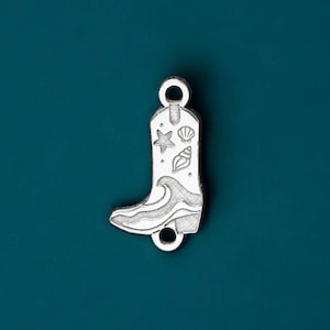 May include: A silver-coloured charm shaped like a cowboy boot. The boot has wave and seashell designs. The charm has loops at the top and bottom for attaching to jewellery. The background is a teal colour.