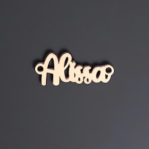 May include: A wooden cutout of the name "Alissa" in a cursive font. The letters are a light tan color, and the cutout is set against a dark gray background. The name is in a flowing script style.
