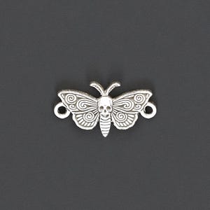 Puede incluir: Un dije plateado con forma de polilla con un diseño de calavera en el cuerpo. Las alas presentan intrincados patrones de remolinos. El dije tiene dos pequeños bucles a cada lado, probablemente para sujetarlo. Fondo gris oscuro.