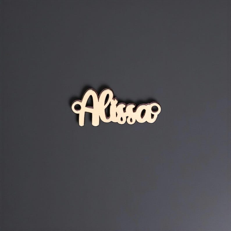 May include: A wooden cutout of the name "Alissa" in a cursive font. The letters are a light tan color, and the cutout is set against a dark gray background. The name is in a flowing script style.