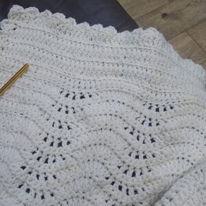 Può includere: Una coperta all'uncinetto bianca con un bordo a festoni. La coperta è realizzata con un punto a trama testurizzata e si trova su una superficie in pelle marrone.