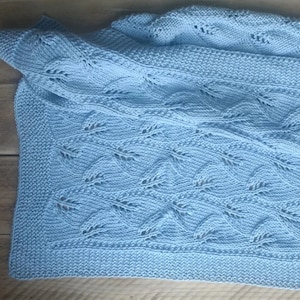 Puede incluir: Una manta de bebé de punto azul claro con un delicado patrón de hojas.