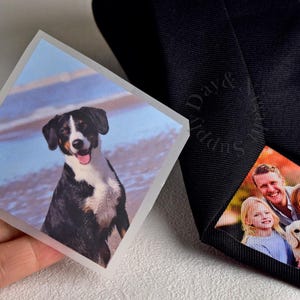 Puede incluir: Una corbata negra con una impresión fotográfica personalizada en la parte posterior. Una foto cuadrada de un perro se sostiene junto a la corbata. El interior de la corbata presenta un retrato familiar. La corbata es un accesorio clásico.