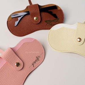 Könnte beinhalten: Drei Leder-Brillenetuis in Braun, Hellgelb und Rosa. Jedes Etui hat einen Klappenverschluss mit einem goldenen Druckknopf. Das braune Etui enthält eine Brille. Das rosa Etui ist mit dem Namen "Jennifer" personalisiert. Das hellgelbe Etui hat ein Blumenlogo.