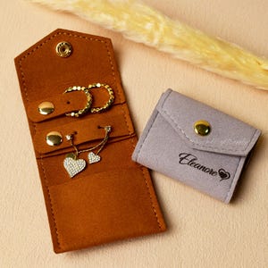 Könnte beinhalten: Ein brauner Schmuck-Organizer aus Wildleder mit goldenen Ohrringen und einer Herz-Anhänger-Halskette. Ein kleines graues Täschchen mit goldenem Druckknopf und dem Namen "Eleanore" liegt daneben. Der Schmuck liegt auf einer hellrosa Oberfläche.