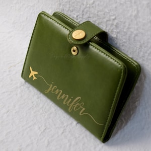 May include: Olive green leather wallet with a gold airplane graphic and the name "Jennifer" in gold script. The wallet has a snap closure with a gold button. The wallet is approximately 10 cm wide and 10 cm tall.