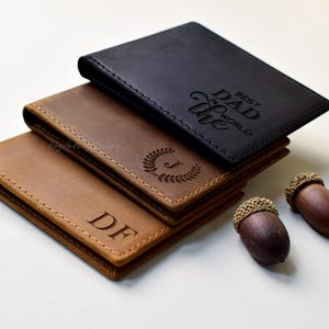Puede incluir: Tres carteras de cuero en tonos marrón y negro apiladas, con dos bellotas a la derecha. La cartera superior es negra y tiene el texto "BEST DAD in the WORLD". Las otras carteras son marrones y tienen iniciales grabadas y una corona de laurel.