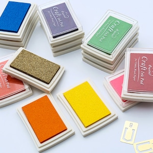 Op de afbeelding: Assortiment van ambachtelijke inktkussens in verschillende kleuren, waaronder blauw, paars, groen, roze, goud, oranje en geel. Elk kussen is gelabeld met "Craft Ink Pad" en "Crystal" met de tekst "For Fabric, Wood Paper".