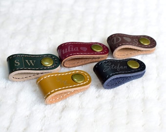 May include: A collection of personalized leather cable organizers in various colors, including green, burgundy, and yellow. Each organizer features a snap closure and is engraved with initials or names. The organizers are approximately 1 inch in length.