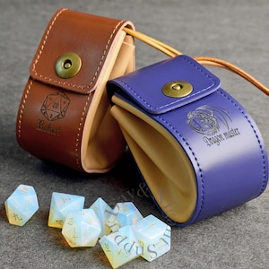 Peut inclure: Deux pochettes en cuir avec fermetures à pression en laiton et intérieurs beige, une marron avec une icône de dé à 20 faces et le nom Robert, une violette avec un motif de dragon et le texte Dragon master, accompagnées de dés blancs translucides.