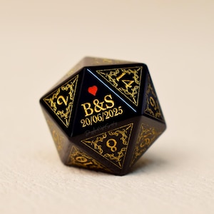 May include: A black and gold D20 die with the initials "B&S", a red heart, and the date "20/06/2025". The die has gold filigree designs around the numbers.