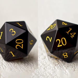 May include: A black twenty-sided die with gold numbers. The die is showing the number 20.
