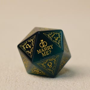 May include: A dark green, twenty-sided die with gold numbers and decorative accents. The text "MARRY ME?" is displayed in gold, along with two interlocking rings. A unique proposal item.