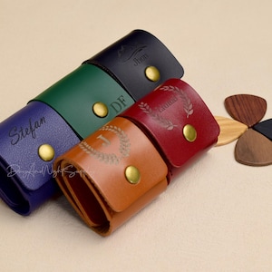May include: Five leather guitar picks cases with different colors and personalized names. The cases are arranged in a row with the names facing the camera. The names are "Stefan", "DF", "Lauren", "Jhon", and a number "1" with a wreath around it.