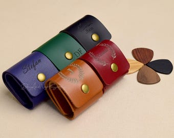 Peut inclure: Cinq étuis en cuir pour médiators de guitare de différentes couleurs, chacun avec un nom personnalisé. Les étuis sont disposés en rangée, les noms étant tournés vers l'appareil photo. Les noms sont "Stefan", "DF", "Lauren", "Jhon" et un numéro "1" avec une couronne autour.
