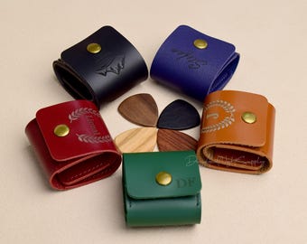 Peut inclure: Plusieurs étuis à médiators en cuir de différentes couleurs : bleu marine, bleu, rouge, marron et vert. Chaque étui est doté d'une fermeture à bouton-pression et d'un bouton doré. Des médiators en bois sont également visibles.