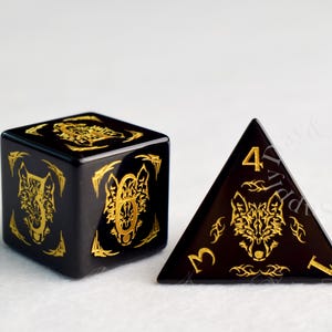 May include: A black six-sided die and a black four-sided die, both featuring gold-colored engravings of a wolf head and tribal-style patterns. The numbers are displayed in a gold font, with the four-sided die showing the number four at the top.