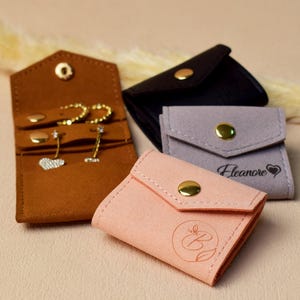 May include: An assortment of small jewelry pouches in various colors, including brown, black, gray, and pink. The pouches are made of soft material and feature gold-colored snap closures. Some pouches have personalized text or designs, and one is open, displaying earrings.