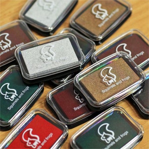 Könnte beinhalten: Eine Sammlung von 10 Stempelkissen in verschiedenen Farben, darunter Silber, Gold, Rot, Grün und Braun. Jedes Kissen hat ein transparentes Plastikgehäuse mit schwarzem Rand und ein weißes Eichhörnchen- und Froschdesign. Der Text "Squirrels and Frogs" ist auf dem Kissen aufgedruckt.