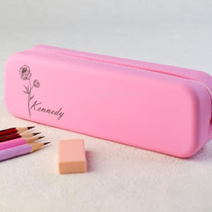May include: A pink pencil case with a zipper closure. The case has a floral design and the name "Kennedy" printed on it. There are also several pencils and an eraser next to the case.