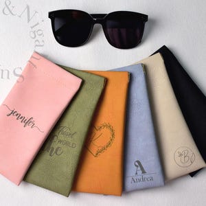 May include: Black sunglasses rest above a collection of colorful eyeglass cases. The cases are in pink, olive green, orange, light blue, beige, and black. Each case has a unique design or name printed on it.