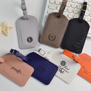 May include: A selection of personalised luggage tags in shades of grey, brown, black, pink, blue, white, and orange. Each tag has a buckle strap and is engraved with initials, names, or travel-themed designs. A keyboard and decorative items are in the background.