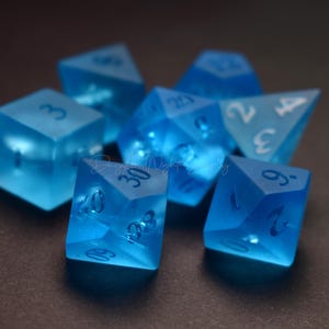 Puede incluir: Un conjunto de siete dados azules con números blancos. Los dados están hechos de un material translúcido, y los números son visibles a través de los dados.