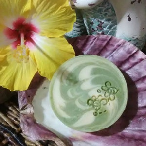 Puede incluir: Una pastilla de jabón de color verde y blanco con un diseño de abeja. El jabón está sobre una concha de mar con un patrón morado y blanco.
