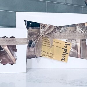 Puede incluir: Tres cajas de regalo blancas con dise&ntilde;os recortados, atadas con cintas y lazos color beige. Cada caja contiene art&iacute;culos visibles a trav&eacute;s de los recortes e incluye una etiqueta con un nombre y la frase "&iquest;Quieres ser mi dama de honor?".