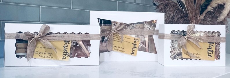 Puede incluir: Tres cajas de regalo blancas con dise&ntilde;os recortados, atadas con cintas y lazos color beige. Cada caja contiene art&iacute;culos visibles a trav&eacute;s de los recortes e incluye una etiqueta con un nombre y la frase "&iquest;Quieres ser mi dama de honor?".