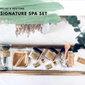 Puede incluir: Un Signature Spa Set con un rodillo de piedra de jade, una cuchara de madera, tarros de sales de ba&ntilde;o y mascarillas faciales, y aceites esenciales. El set incluye una bandeja de madera y un pa&ntilde;o de algod&oacute;n. El texto "RELAX & RESTORE SIGNATURE SPA SET" es visible.