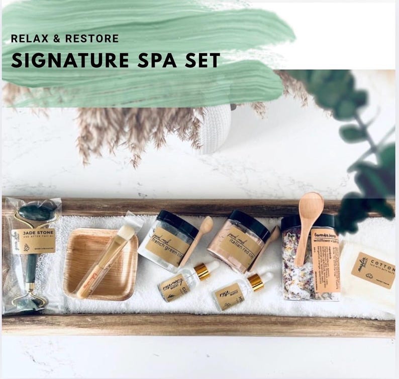 Puede incluir: Un Signature Spa Set con un rodillo de piedra de jade, una cuchara de madera, tarros de sales de ba&ntilde;o y mascarillas faciales, y aceites esenciales. El set incluye una bandeja de madera y un pa&ntilde;o de algod&oacute;n. El texto "RELAX & RESTORE SIGNATURE SPA SET" es visible.