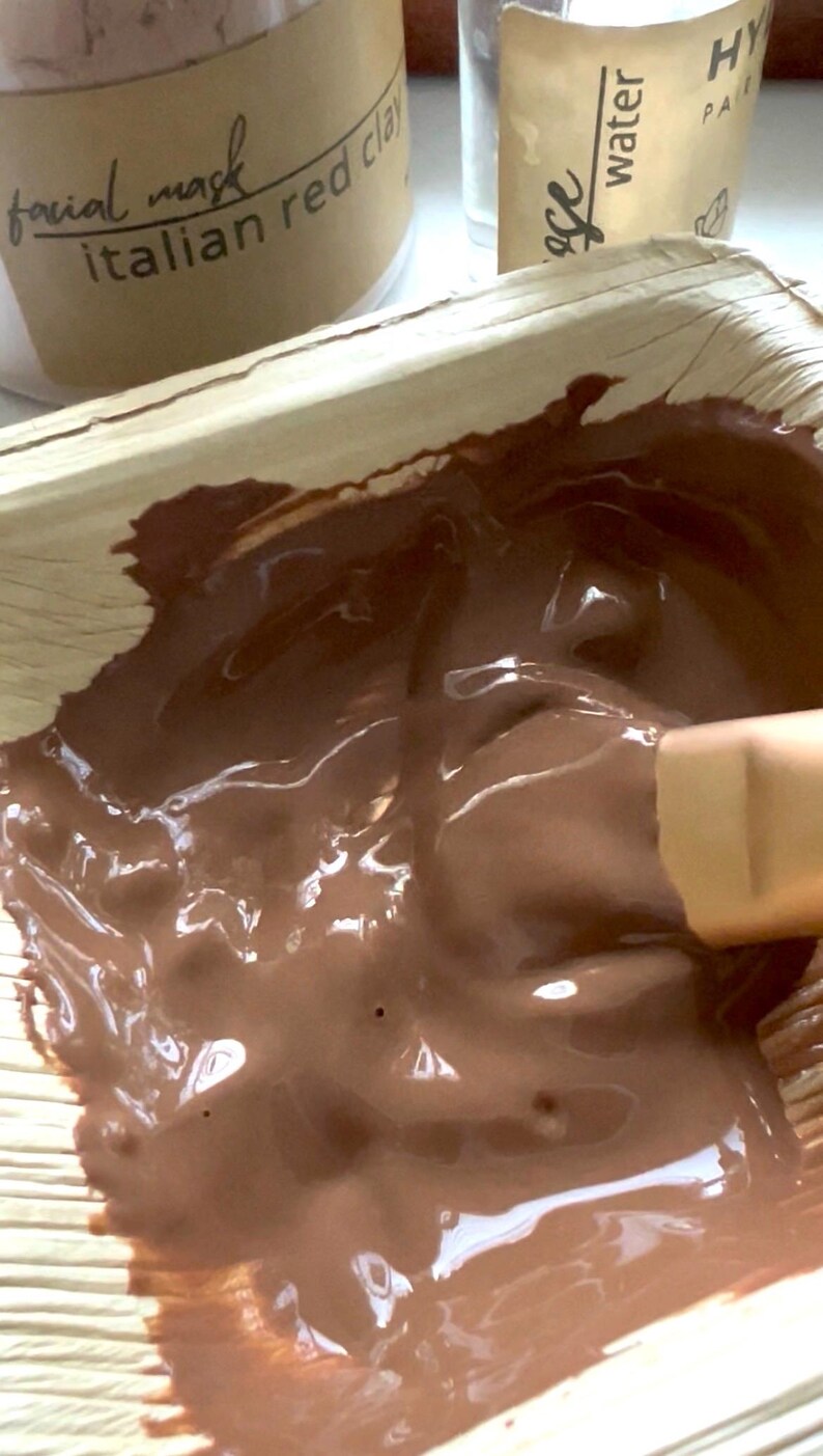 Puede incluir: Primer plano de una mascarilla facial marr&oacute;n, probablemente arcilla roja italiana, mezclada en un cuenco natural con forma de hoja. Se ve un peque&ntilde;o pincel, junto con un tarro etiquetado como "facial mask" y "Italian red clay".