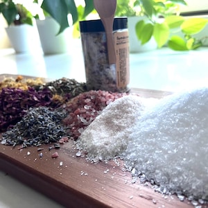 Puede incluir: Una tabla de madera presenta varios tipos de sal y hierbas secas. Montones de sal rosa, blanca y gruesa se disponen junto a lavanda seca, p&eacute;talos de rosa y otros productos bot&aacute;nicos. Un tarro de sal infusionada est&aacute; de pie.