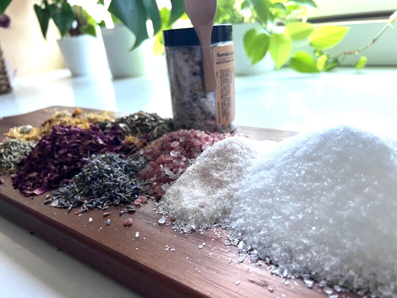 Puede incluir: Una tabla de madera presenta varios tipos de sal y hierbas secas. Montones de sal rosa, blanca y gruesa se disponen junto a lavanda seca, p&eacute;talos de rosa y otros productos bot&aacute;nicos. Un tarro de sal infusionada est&aacute; de pie.