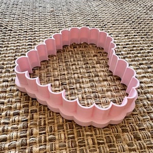 May include: A pink plastic cookie cutter shaped like a scalloped heart. The cutter is on a brown woven surface.