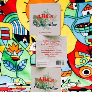 May include: Two stacked children's books titled "ABCs of El Salvador" with a colourful illustration of a sun, flowers, and houses on the cover. The back of the top book has text in both English and Spanish.
