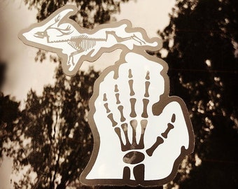 May include: A white vinyl decal shaped like the state of Michigan, featuring a black skeletal hand graphic in the lower peninsula and a black skeletal jackalope graphic in the upper peninsula, with a dark outline against a reflective surface.