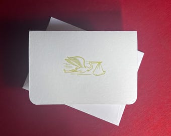 May include: A white card with a gold stork carrying a bundle, resting on a white envelope. The card has rounded corners and a textured surface. The background is a deep red color.